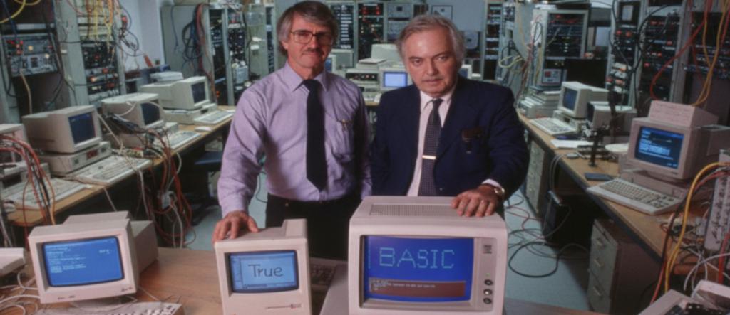 Fotografia em um ambiente de laboratório, repleta de cabos e equipamentos de informática antigos, mostrando dois homens em pé, Thomas Kurtz e John Kemeny, criadores da linguagem BASIC, com suas mãos apoiadas sobre monitores de tubo de raios catódicos (CRT) de computadores de mesa, transmitindo um clima de pesquisa tecnológica dos anos 1990.